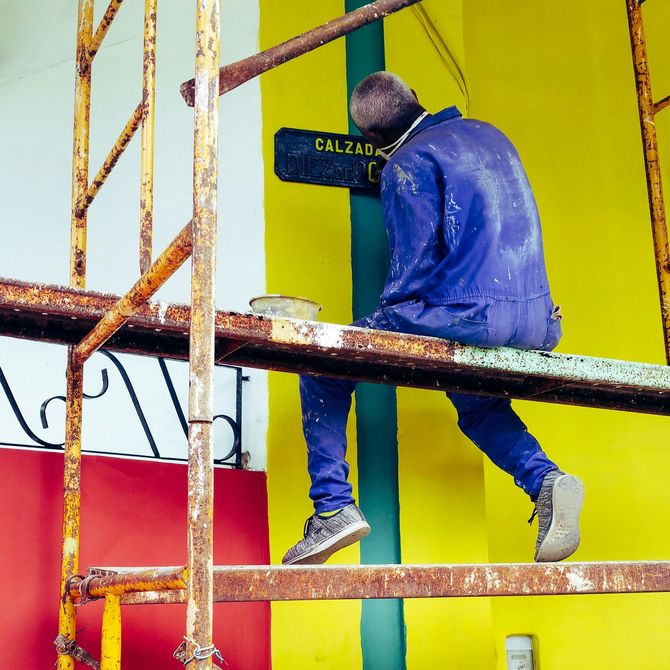 A person in work clothes stands on a stepladder to paint the blue trim of a tall door on a light-colored building.