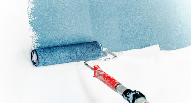 Two workers paint the facade of a white building, one standing on scaffolding and one on the ground near a yellow sign.