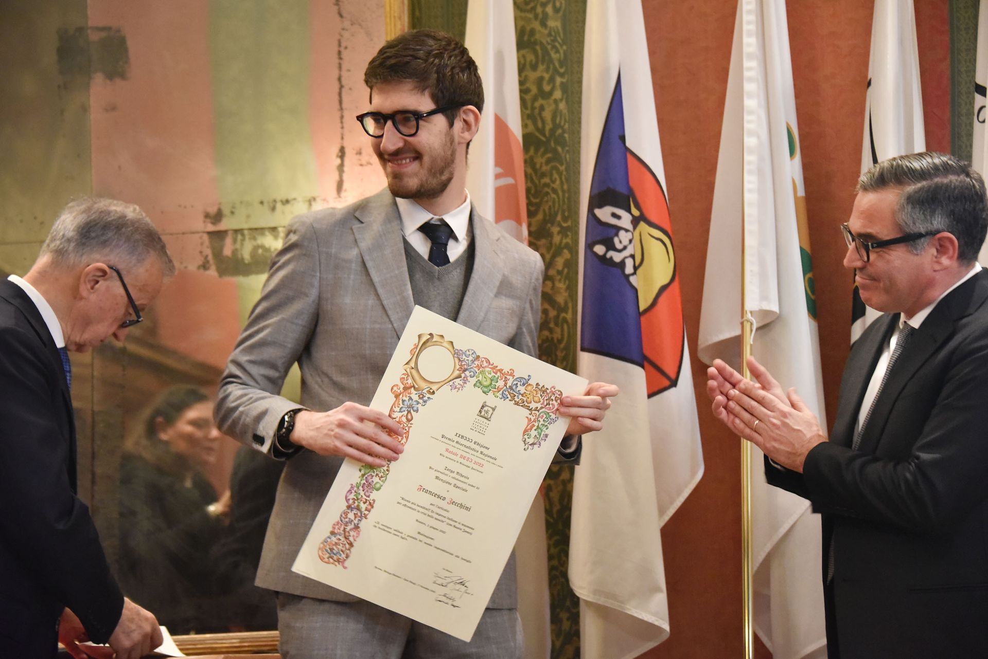 Un uomo in giacca e cravatta tiene in mano un certificato mentre altri due uomini applaudono.