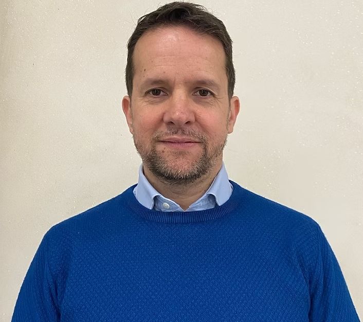 Un uomo calvo con una camicia a quadri sorride alla telecamera.