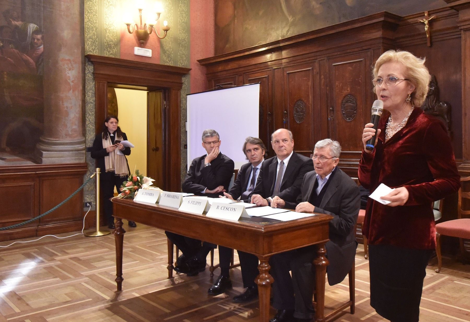 Una donna tiene in mano un microfono davanti a un gruppo di persone sedute a un tavolo.