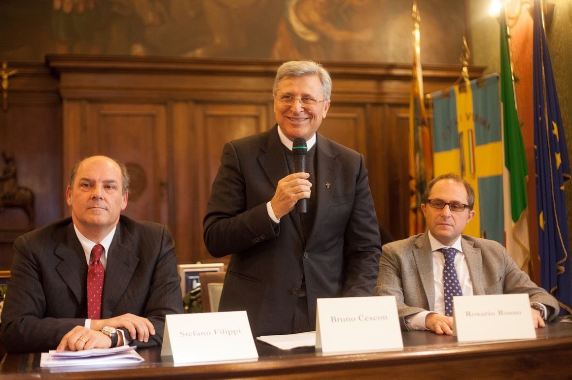 Un uomo in giacca e cravatta parla al microfono mentre altri due uomini sono seduti a un tavolo.