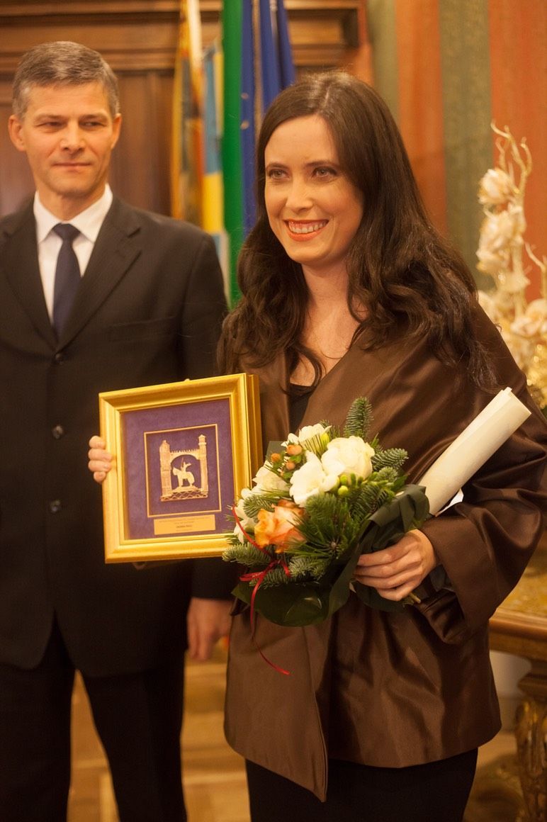 Una donna con una giacca marrone tiene in mano un mazzo di fiori e un'immagine incorniciata.