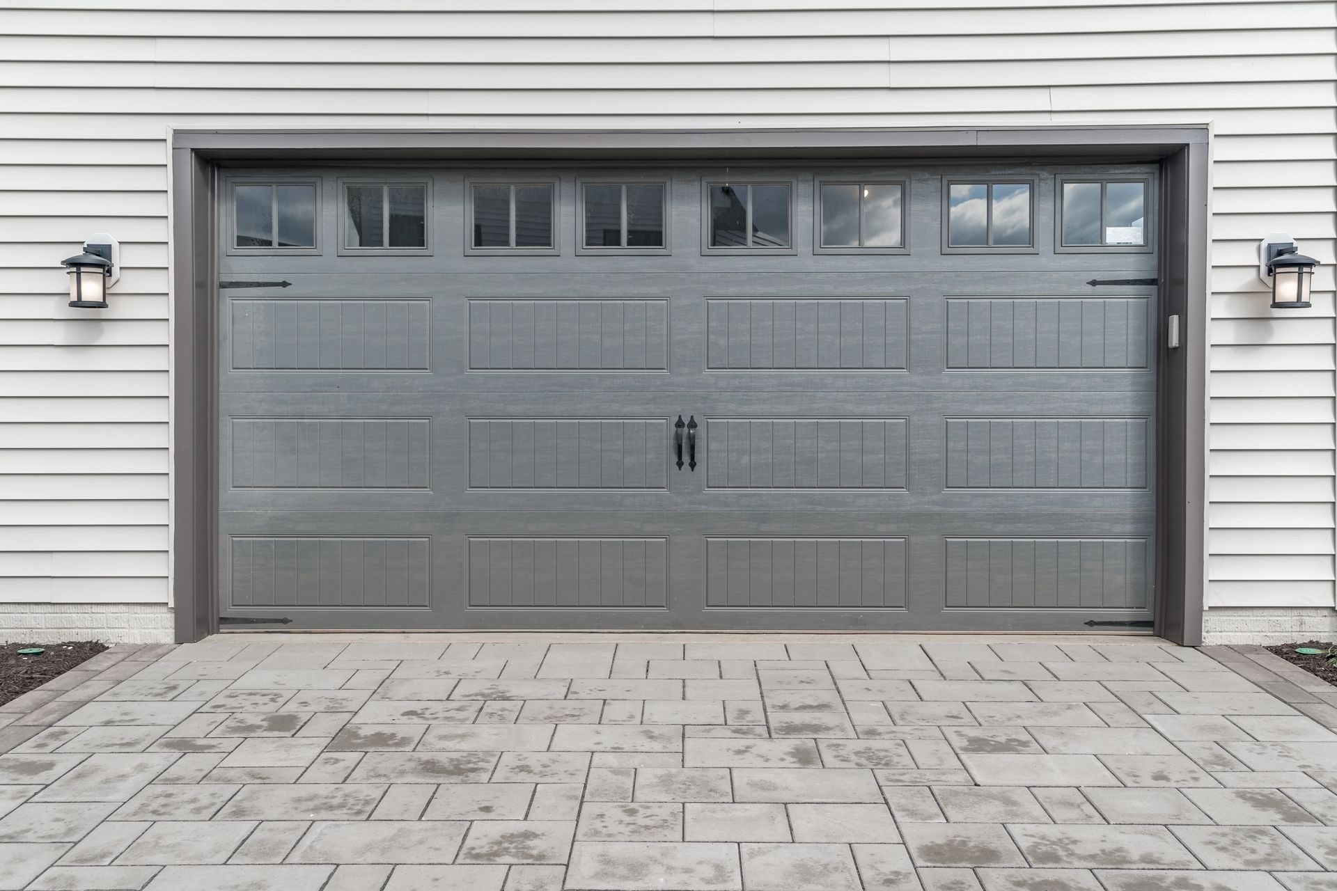 Porte de garage grise avec fenêtres, flanquée de lumières, sur fond de revêtement blanc. Allée en briques.
