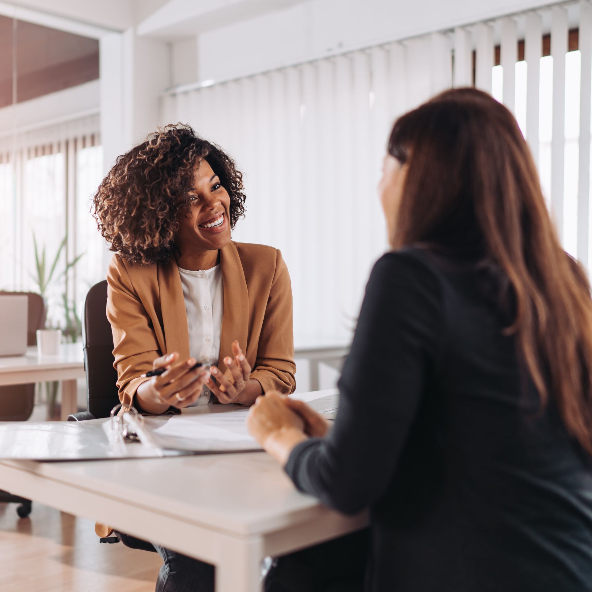 Woman in blazer smiles, gestures while speaking to another woman at a desk in an office setting.