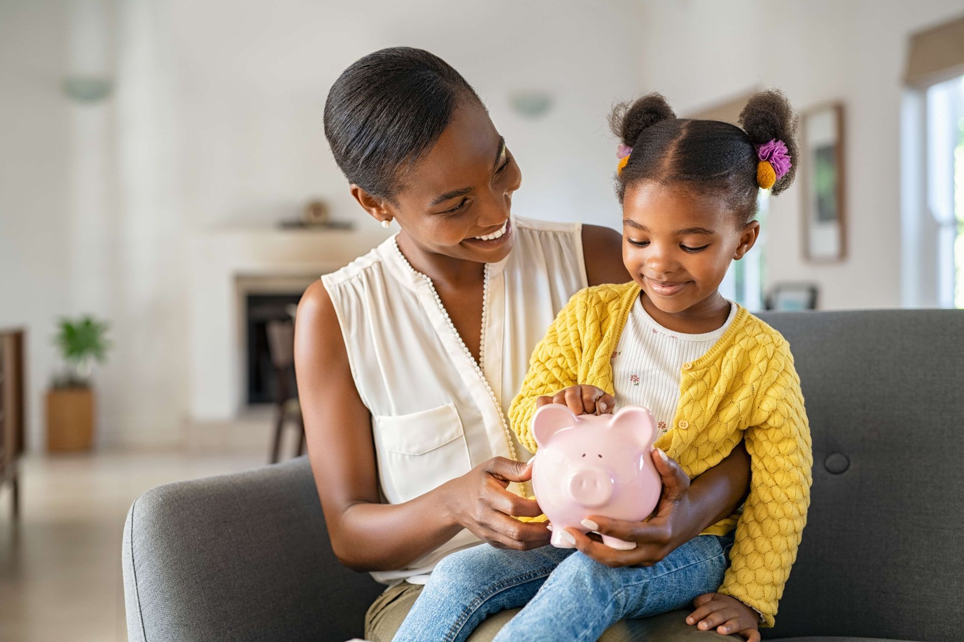 Woman and girl putting money in a pink piggy bank, smiling in a living room.