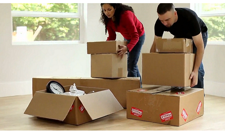 Couple packing moving boxes in a room with large windows. Woman in red and man in black lift boxes, ready to move.