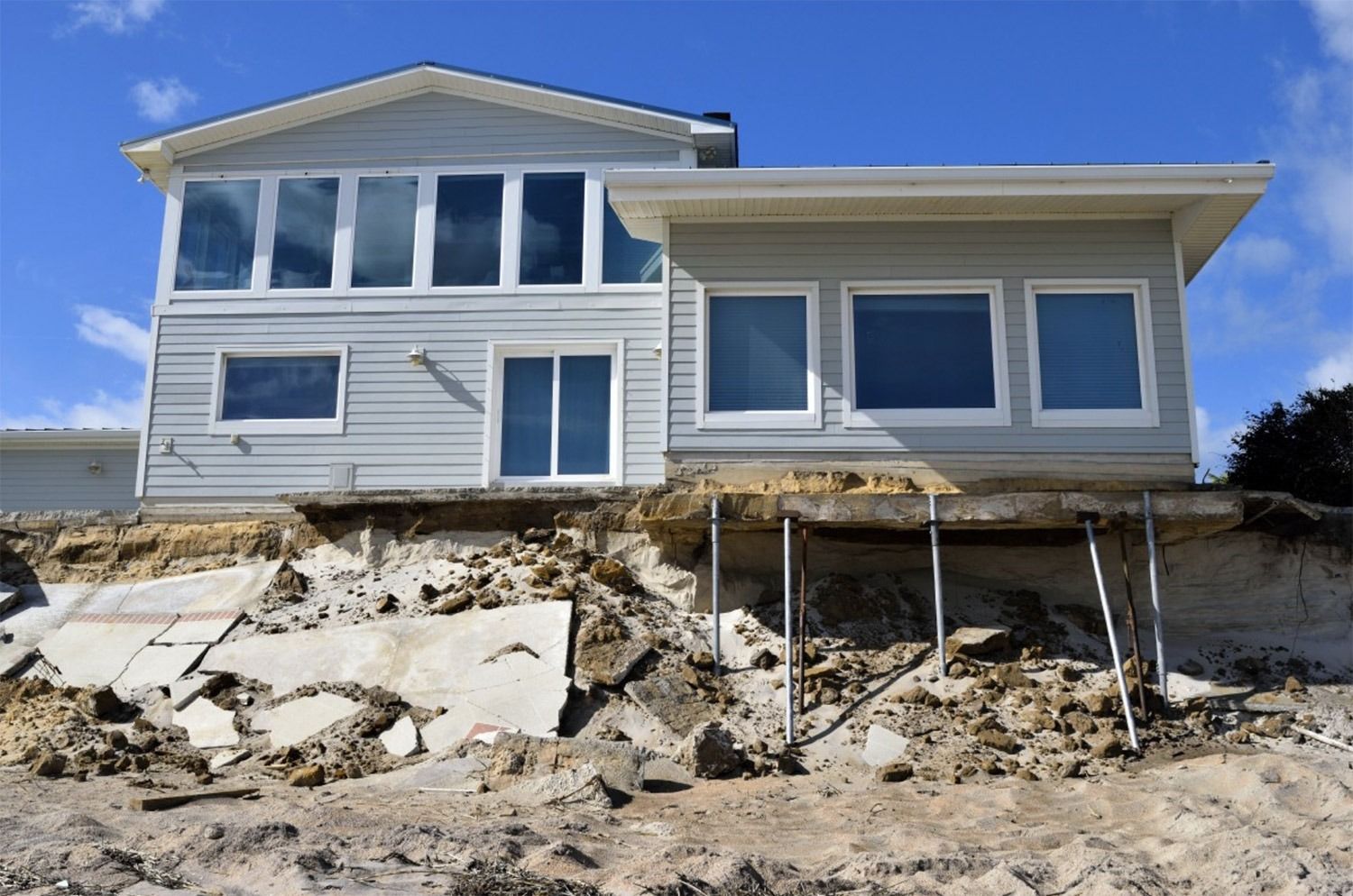 A two-story house with damaged foundation, propped up by metal supports, sits on eroded land.