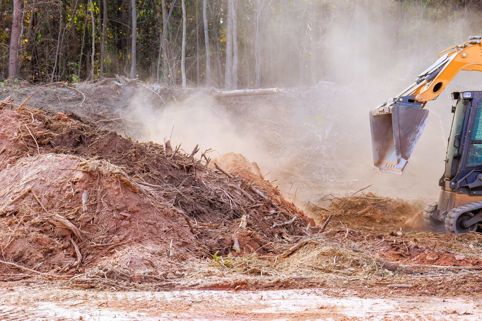 Excavator clearing a large mulch pile in a dusty wooded area