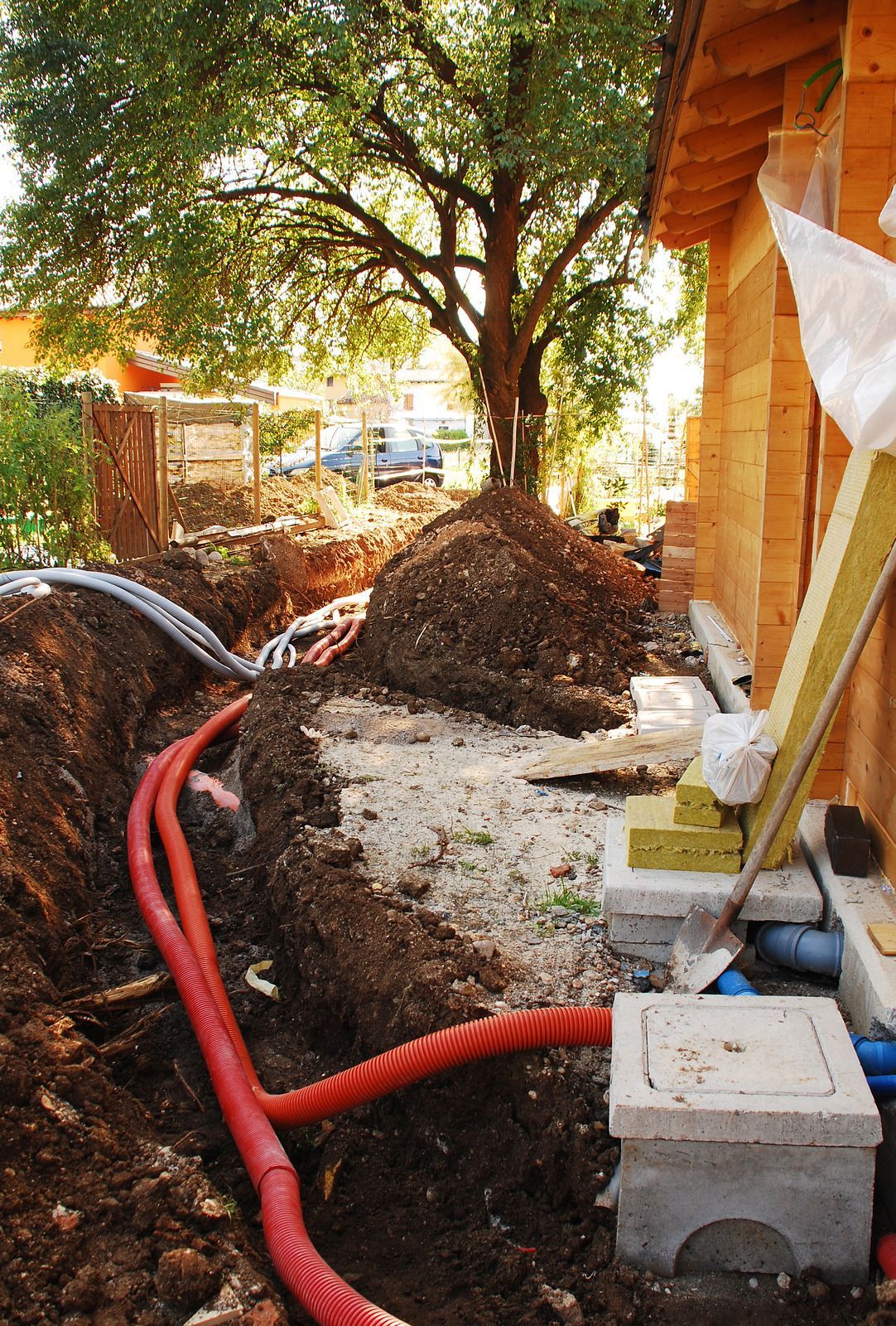 Backyard trench with red hose and dirt pile beside a house foundation under a tree