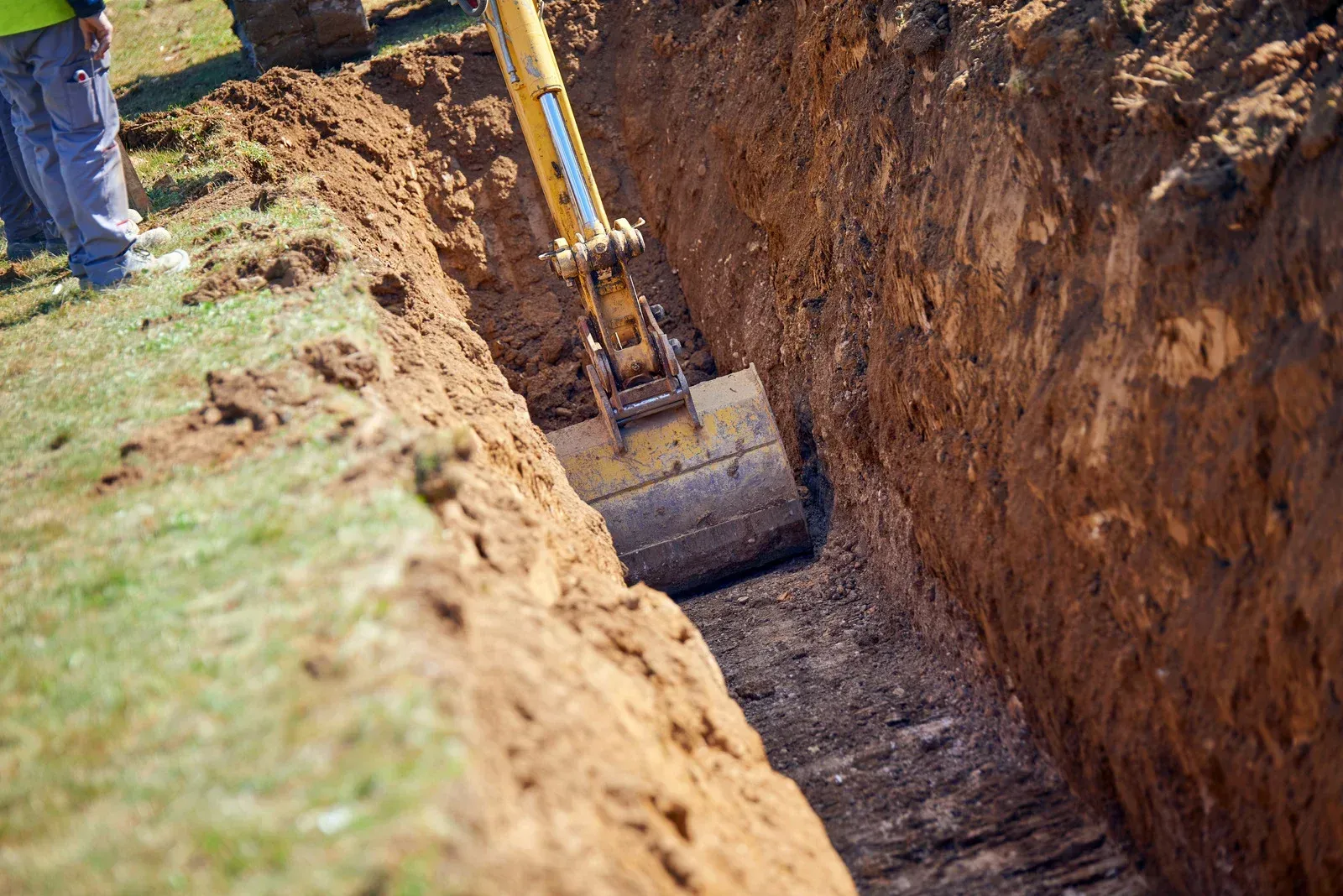 Excavator digging a trench beside a grassy field, with a worker standing nearby.