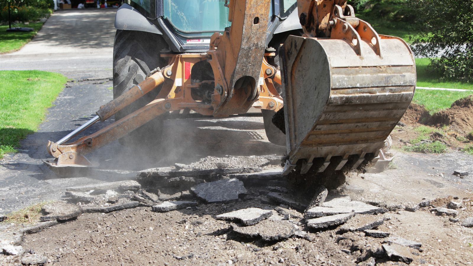 Excavator bucket breaking up a concrete driveway, with dust and debris on the ground.