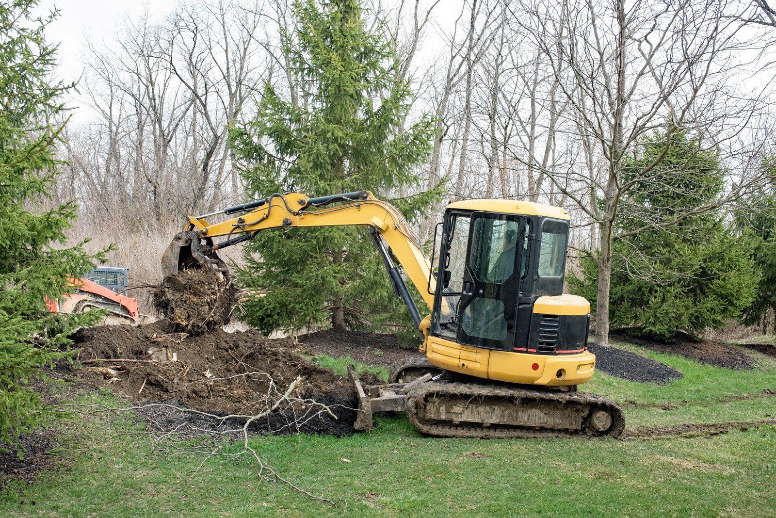 Yellow excavator digging dirt beside trees in a grassy yard