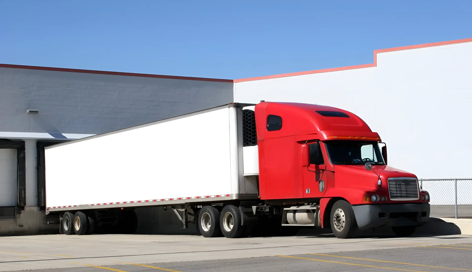 Red semi-truck backed up to a loading dock at a white building under a clear, blue sky.