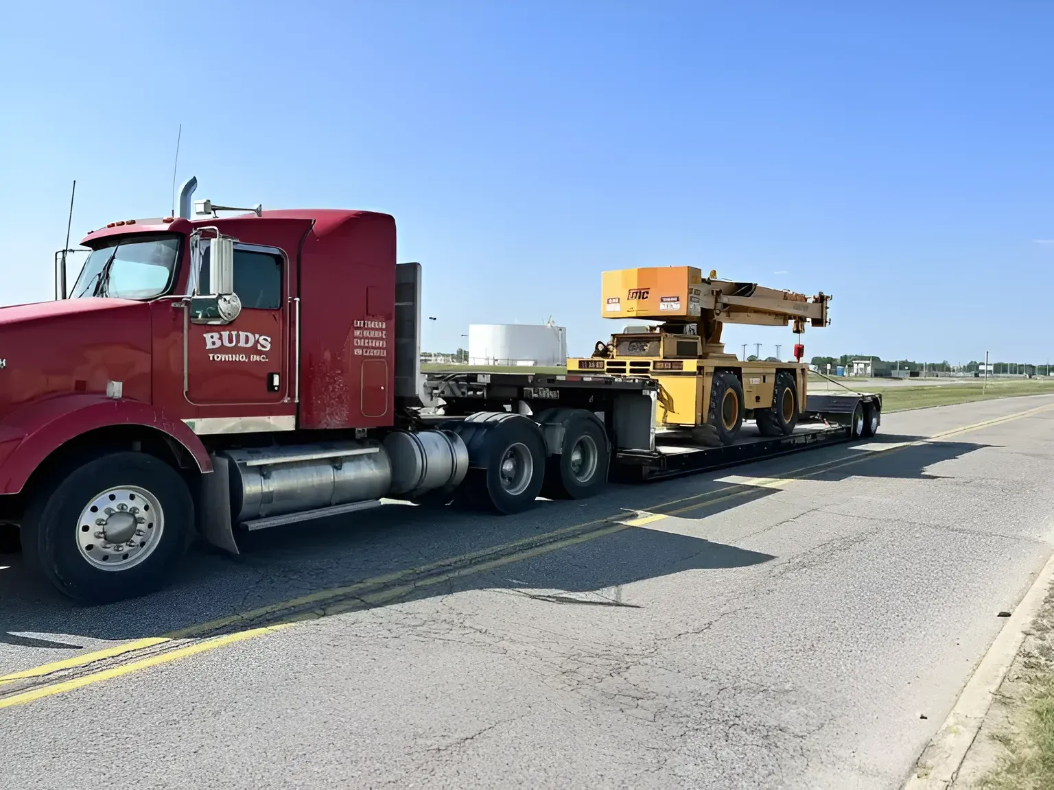 Red semi-truck hauling a yellow crane on a flatbed trailer on a paved road under a blue sky.