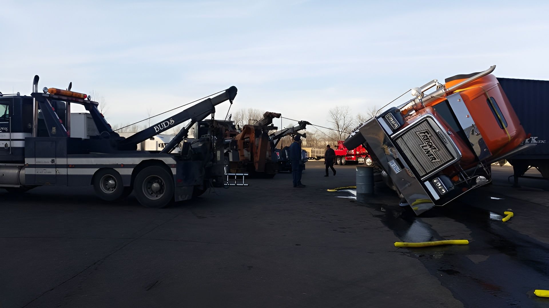 Tow truck lifting an orange semi-truck cab. The cab is tilted. Two other trucks and people are in the paved lot.