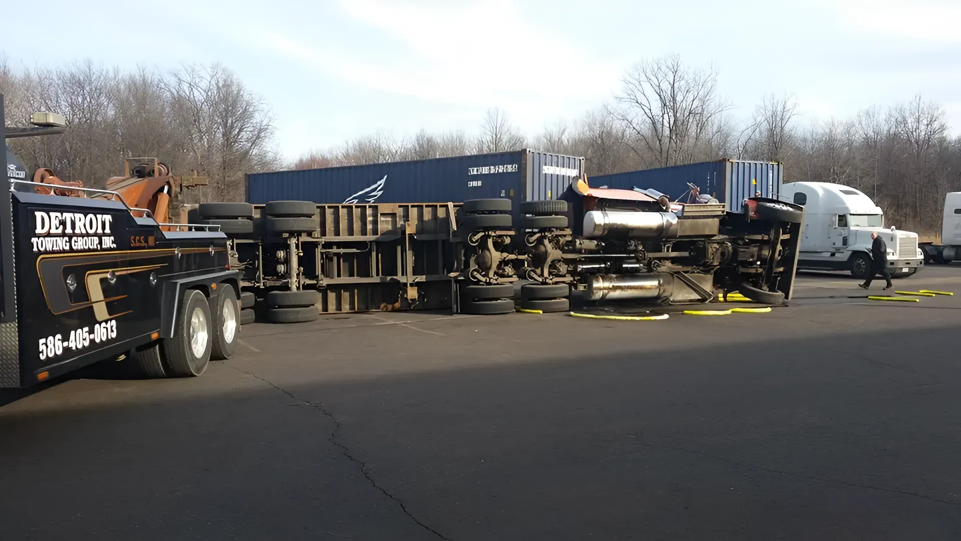 Overturned semi-truck with trailer, Detroit logo on tow truck, blue shipping containers in background.