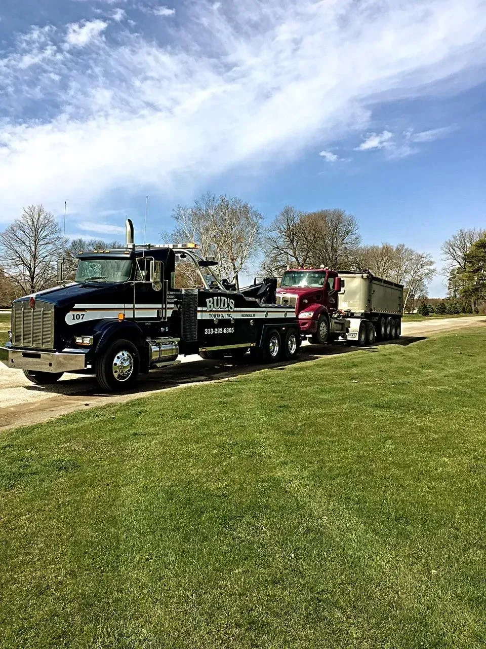 Tow truck towing a semi-truck with a trailer on a grassy area under a partly cloudy sky.