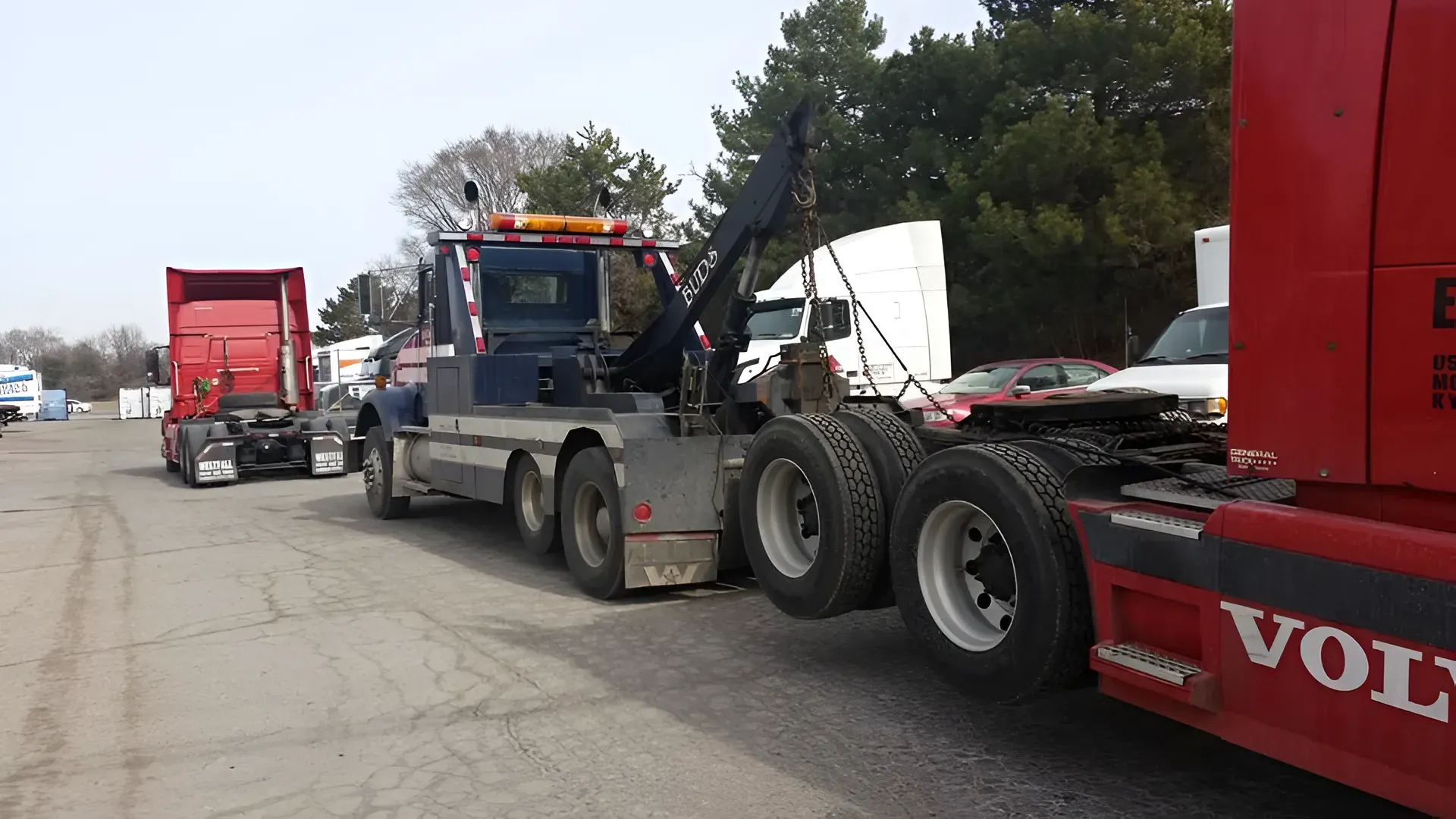 Tow truck hooking up to a red semi-truck. Other trucks are parked in a lot under a blue sky.