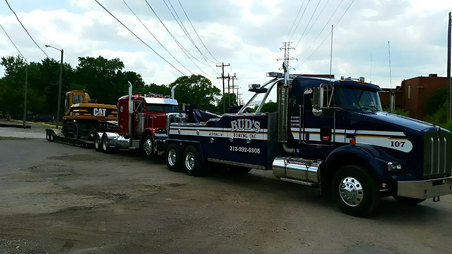 Dark blue tow truck towing a red semi-truck with a yellow bulldozer on a trailer. Cloudy sky.