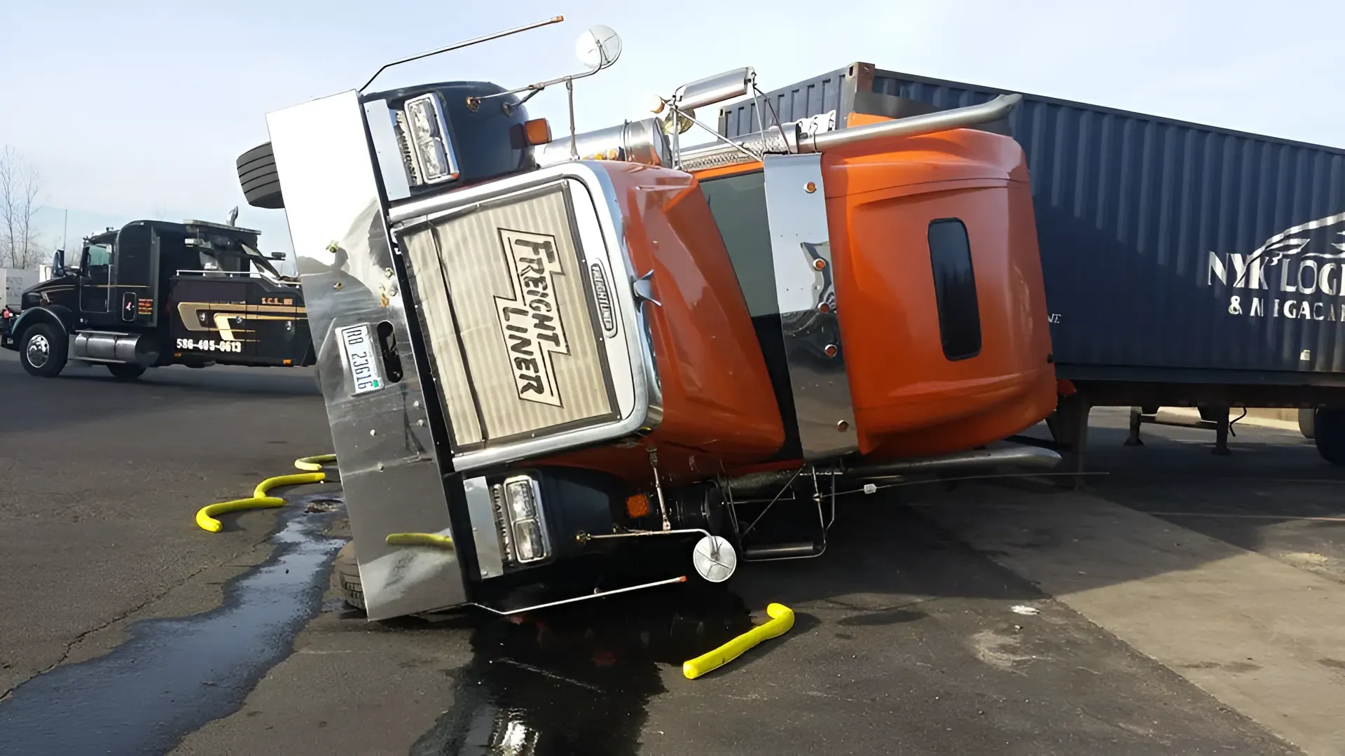 Overturned orange semi-truck with trailer on side, yellow safety devices, tow truck in background.