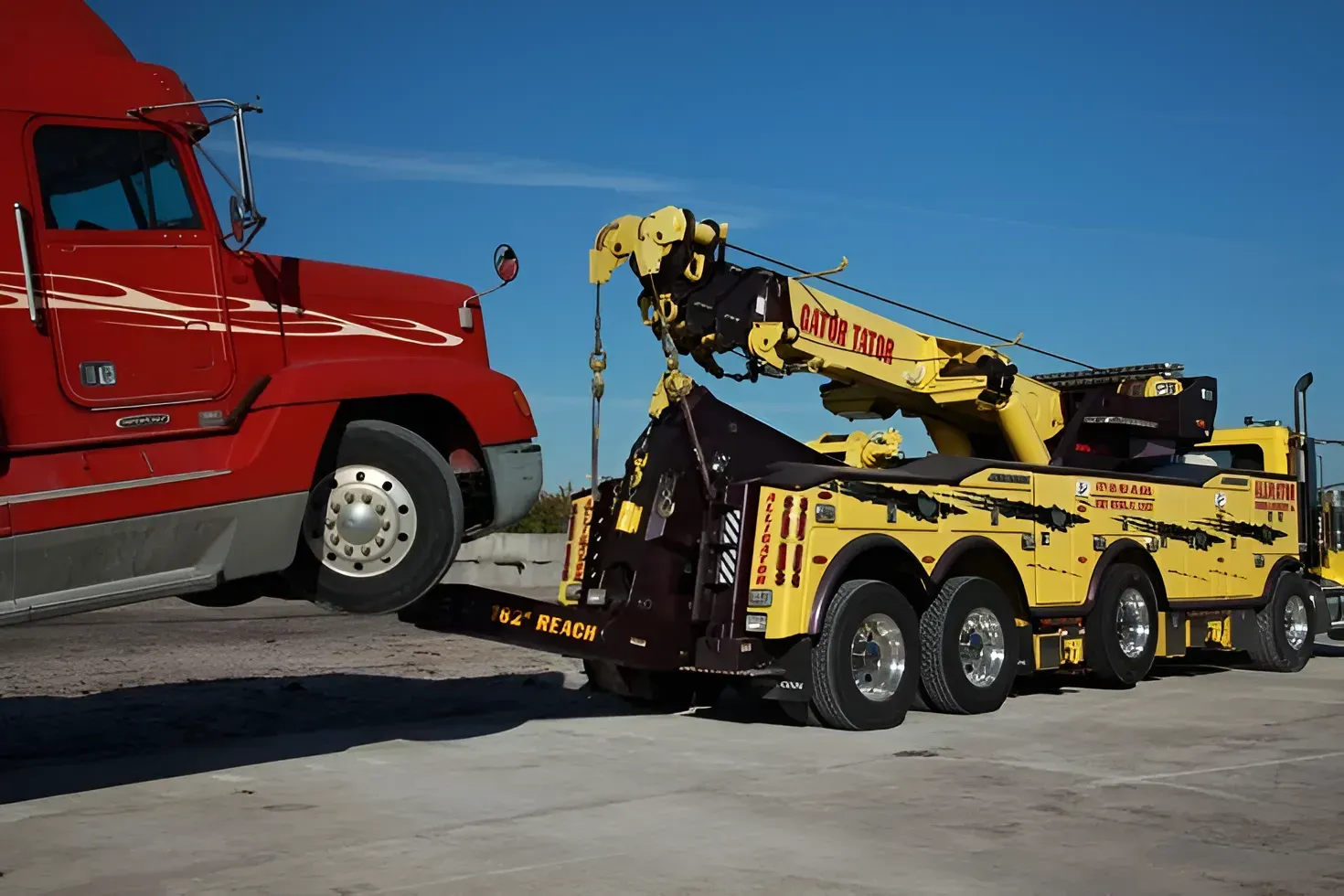 Yellow tow truck towing a large red semi-truck on a concrete surface against a blue sky.