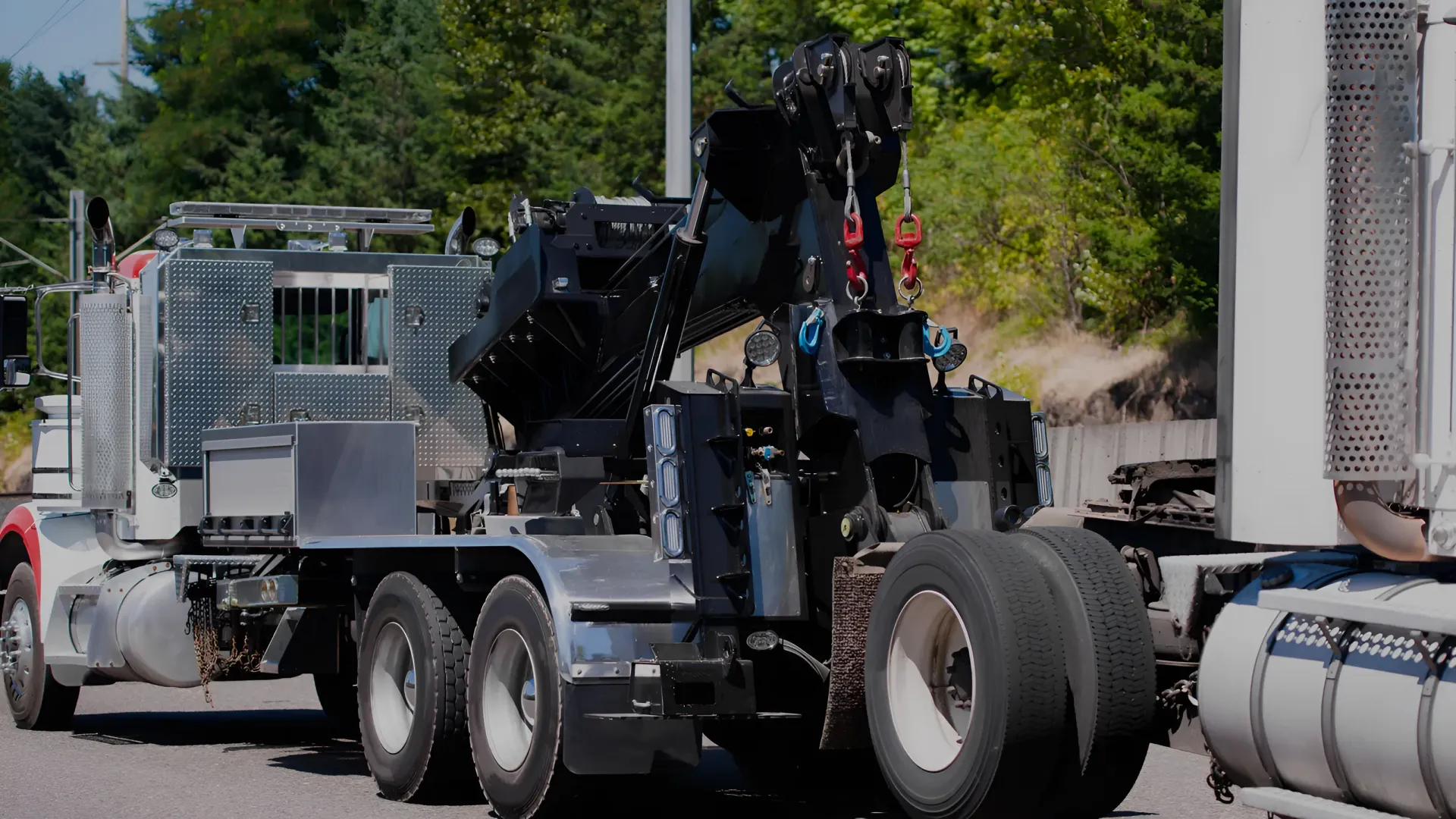 A large black tow truck on a road, hooking up to a white truck's trailer.