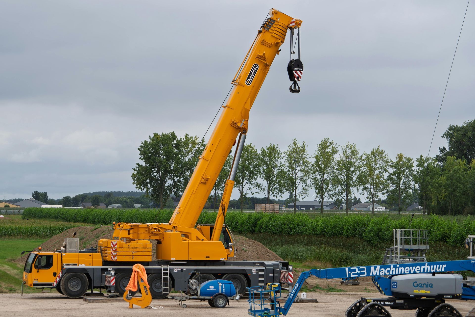 Yellow mobile crane on a construction site with trees and blue lift.