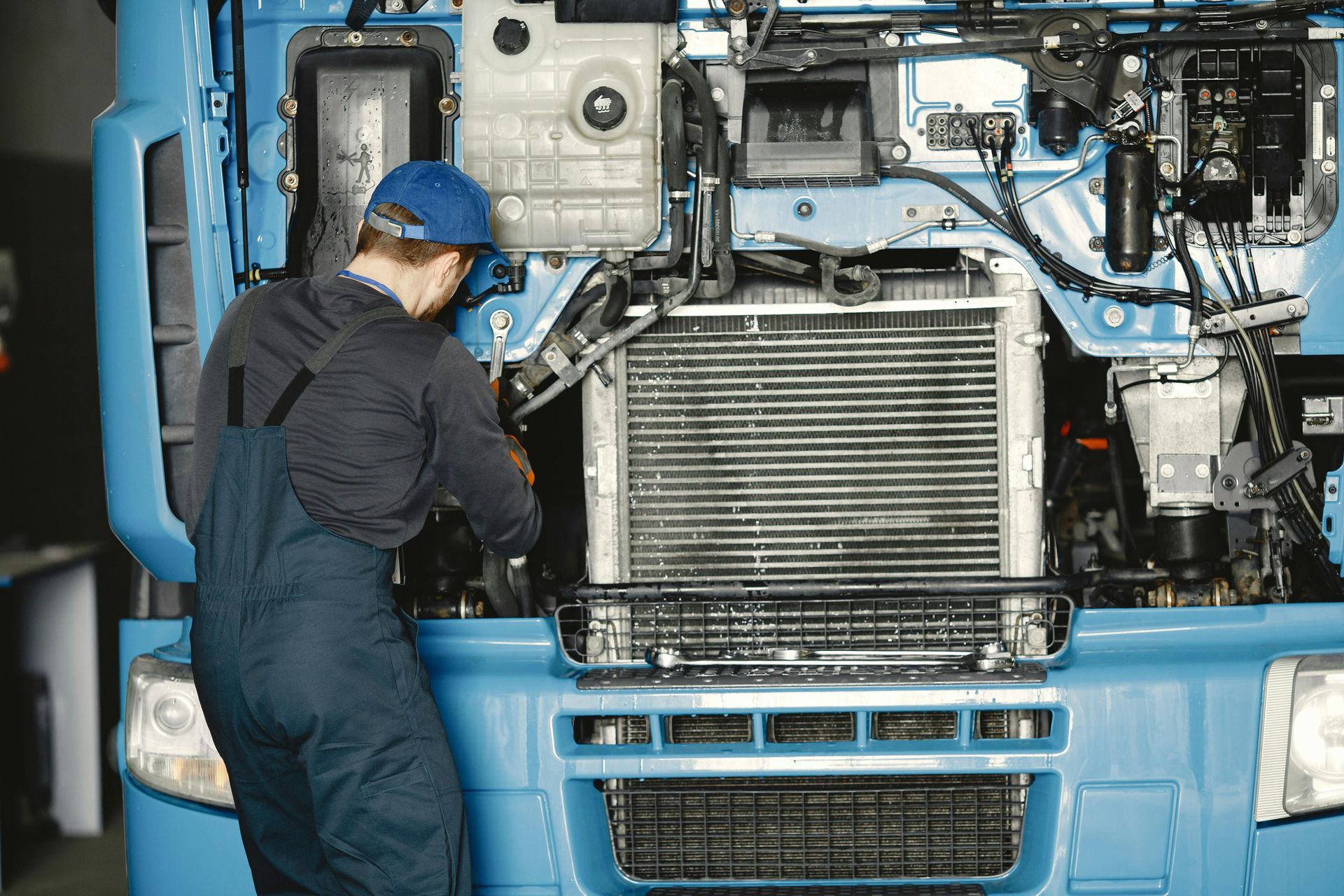 Mechanic in blue overalls working on the front of a blue truck, in a garage.