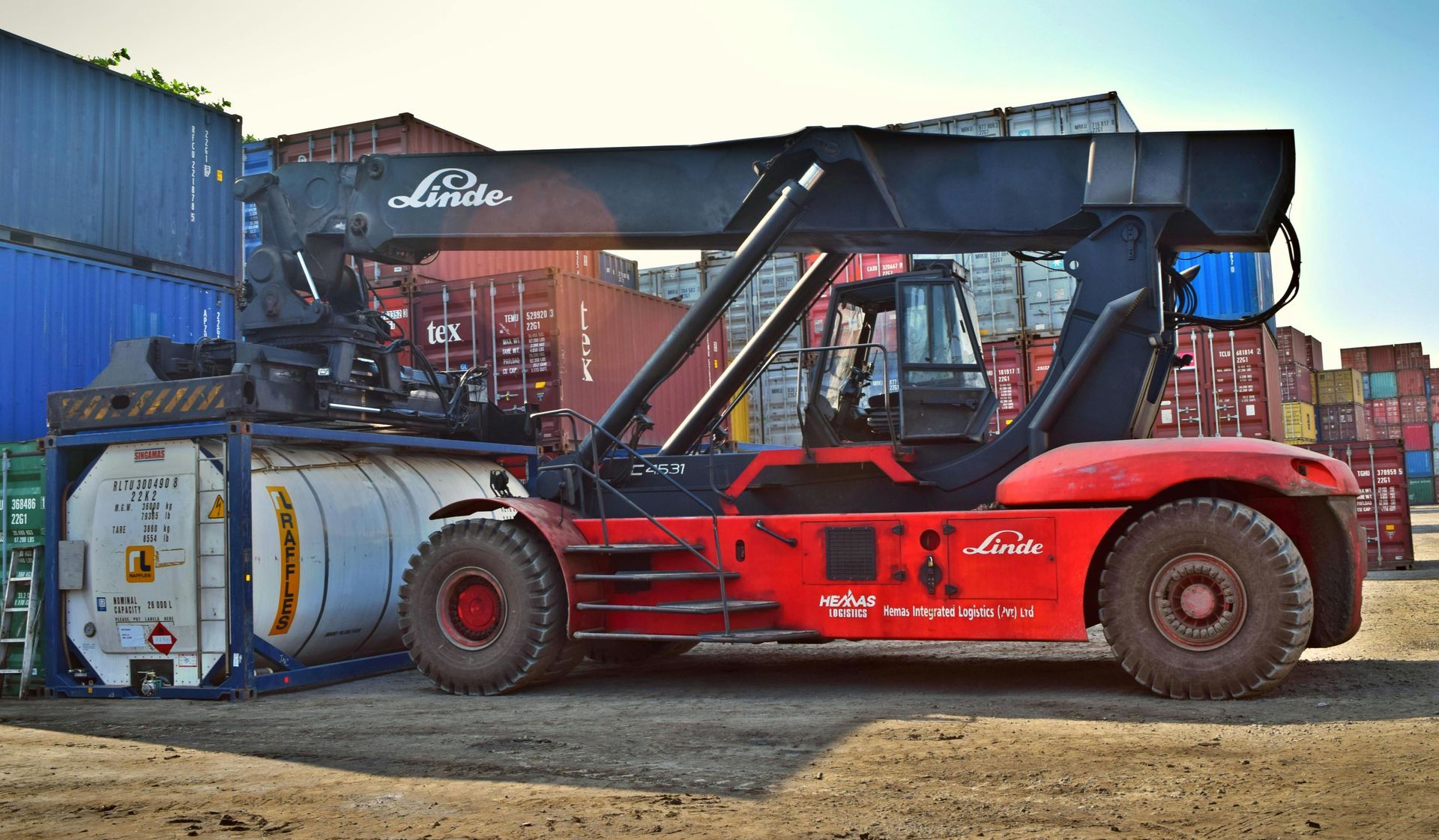 Red Linde reach stacker lifting a white tank container in a shipping yard.