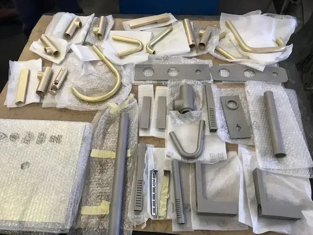 Su un tavolo sono disposti vari componenti metallici di colore bianco e beige, molti dei quali avvolti in imballaggi protettivi.