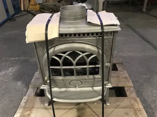 Piccola stufa in ghisa bianca su un pallet di legno, con cinghie nere e condotto metallico sulla parte superiore.
