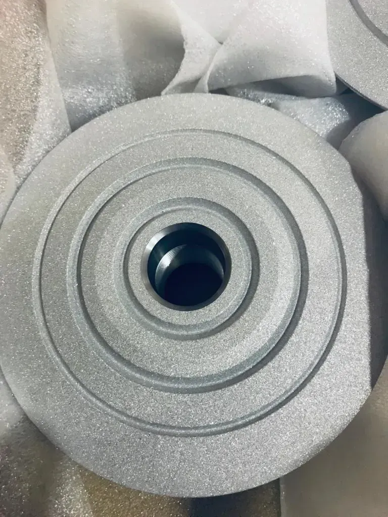 Pezzo metallico circolare grigio con foro centrale, avvolto in un imballaggio di schiuma bianca.