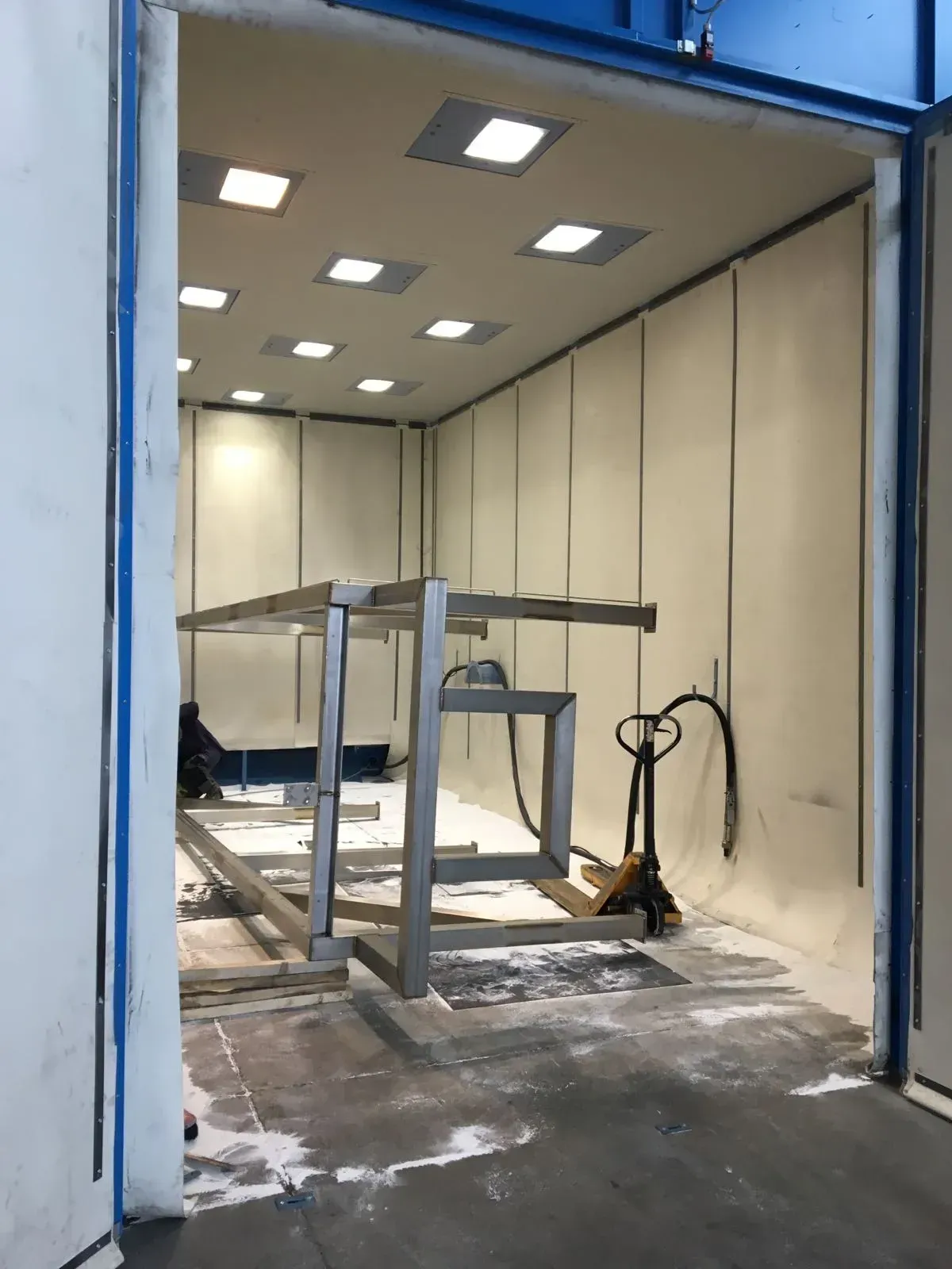 Locale industriale vuoto con tavoli da lavoro in metallo, pareti bianche e pavimento coperto di polvere o detriti.