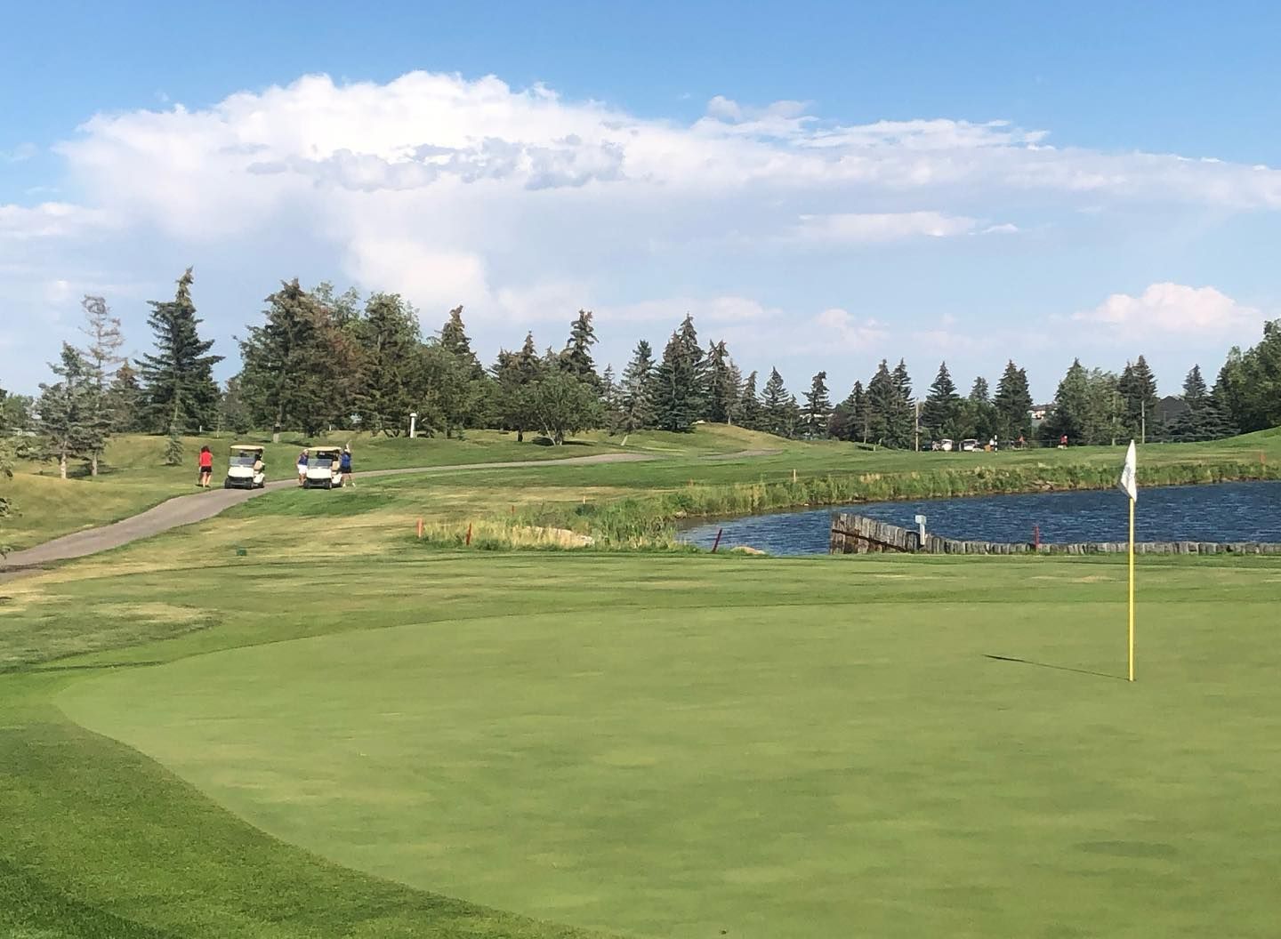 Green golf course with a flag, pond, and golf carts under a blue sky.