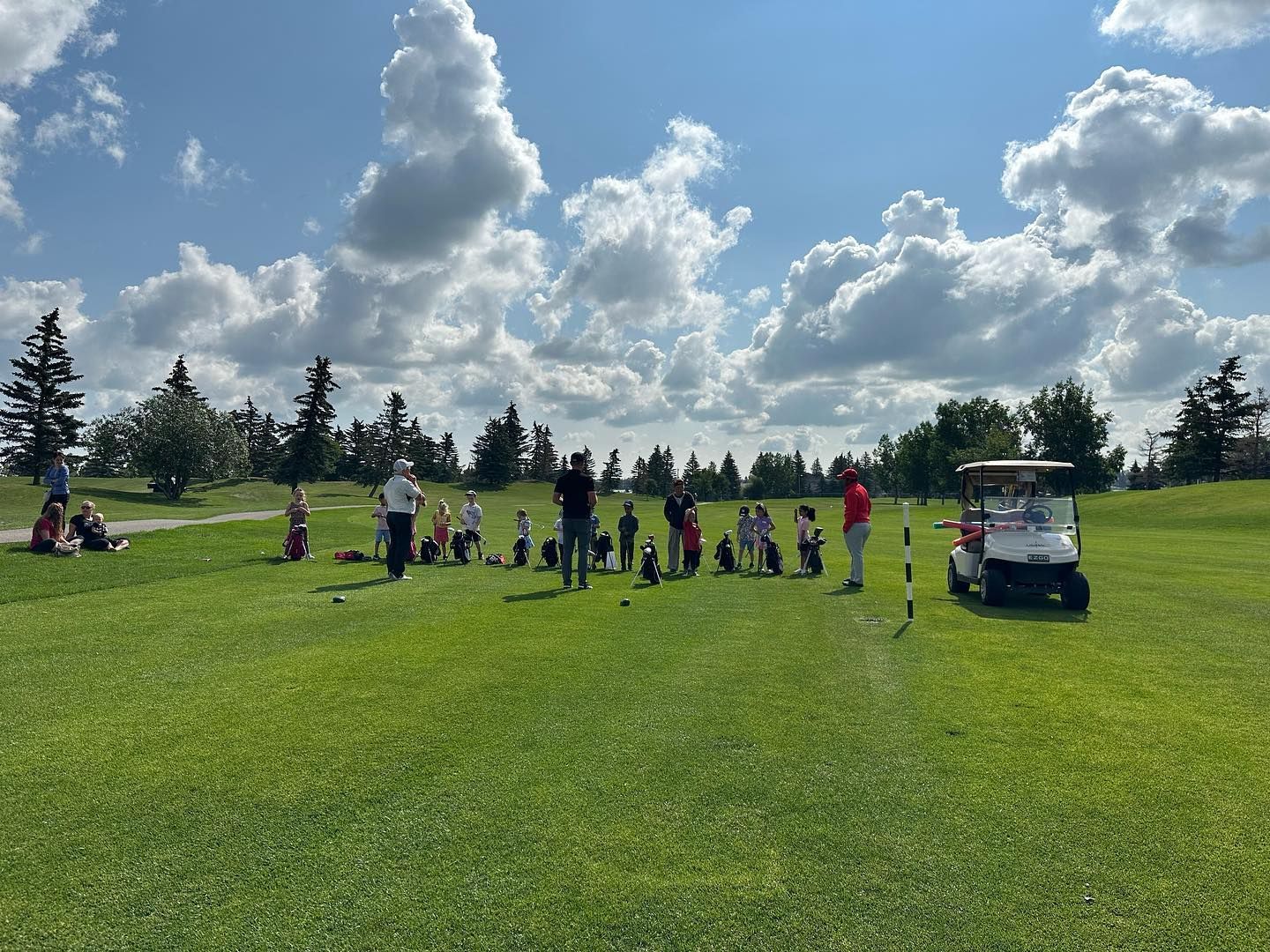Group of kids on a green golf course, with instructors and a golf cart, under a cloudy sky.