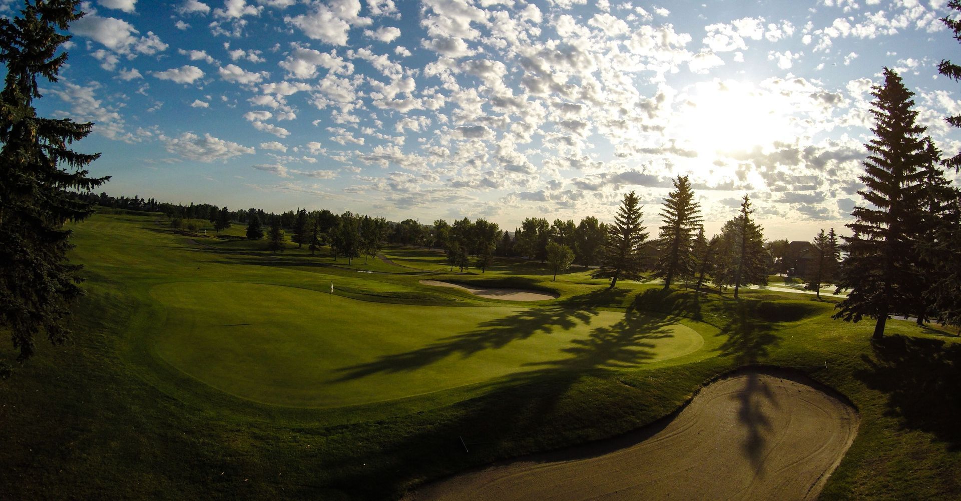 Green golf course under a blue sky with white clouds, trees, and sunlight.