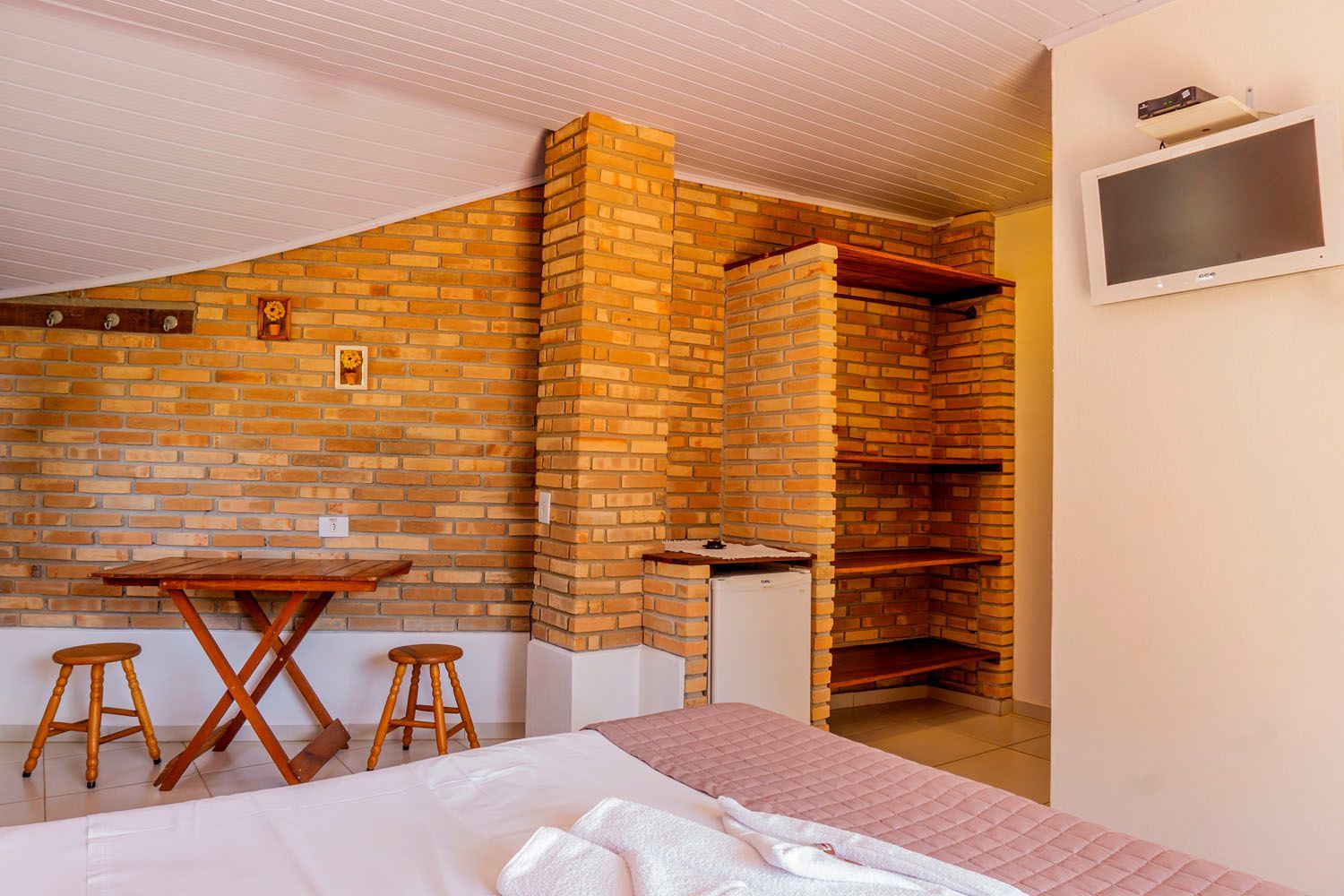 Um quarto com uma cama, mesa, bancos e uma televisão.