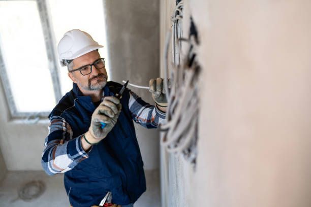 An electrician in a hard hat and work vest uses wire cutters on electrical cables protruding from a wall.