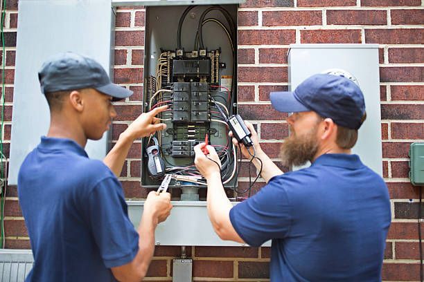 Two electricians in blue uniforms test a residential electrical panel mounted on an outdoor brick wall.