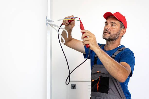 An electrician in a red cap and grey overalls uses a voltage tester on wires coming out of a white wall.