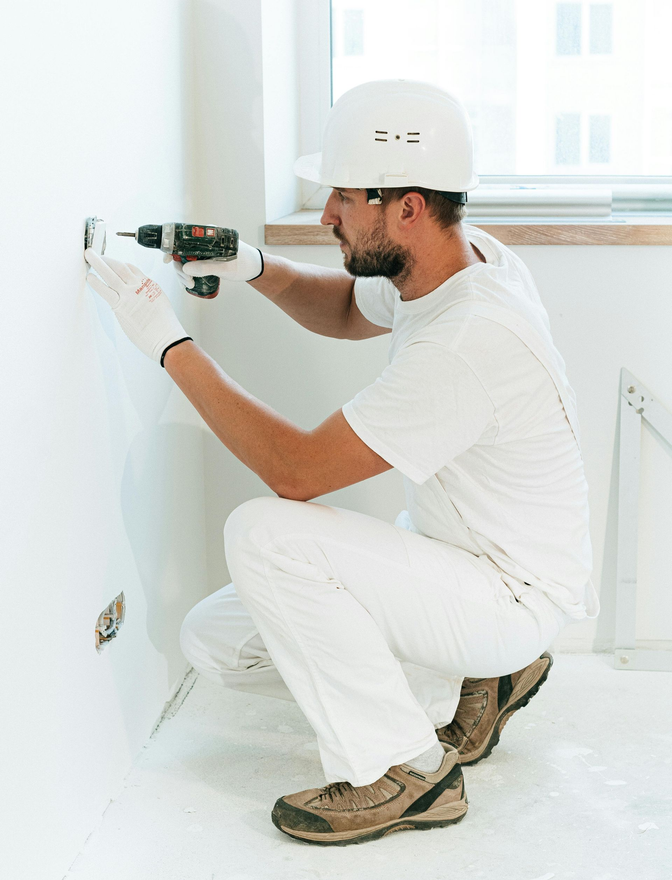 A person wearing a white hard hat and work clothes kneels while using a power drill on a white wall in a room.