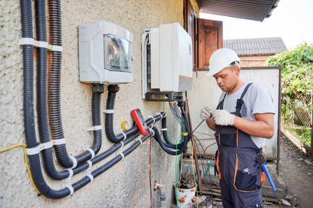 An electrician in a hard hat and work coveralls installs electrical wiring for outdoor equipment on a stucco wall.