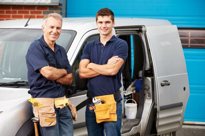Two workers in blue polo shirts and tool belts stand with arms crossed in front of a silver work van.