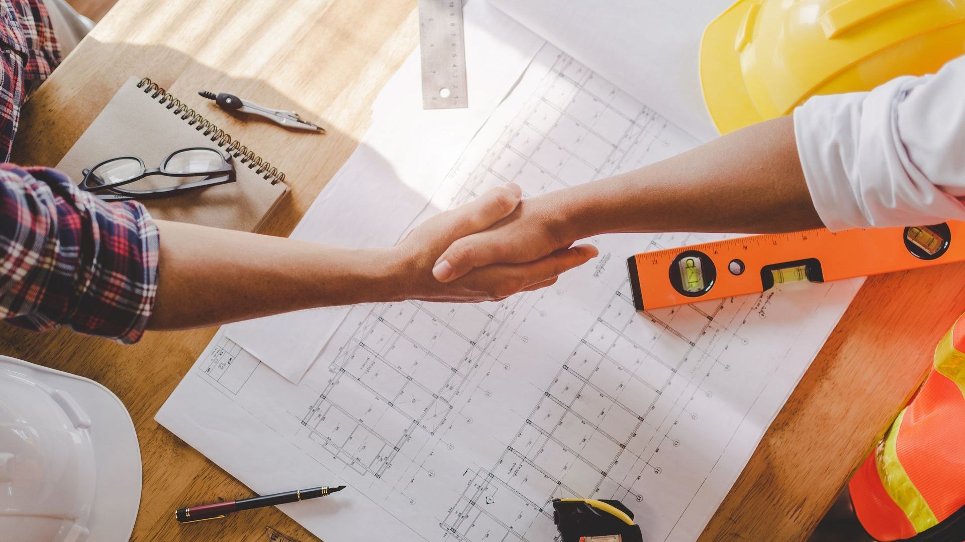 Two construction workers are shaking hands over a blueprint.