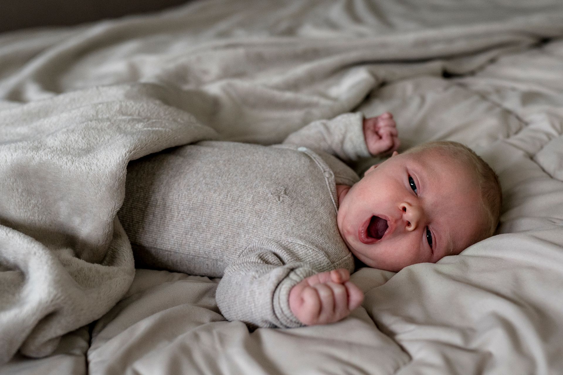 De baby gaapt en rekt zich uit, liggend op een bed bedekt met een grijze deken.