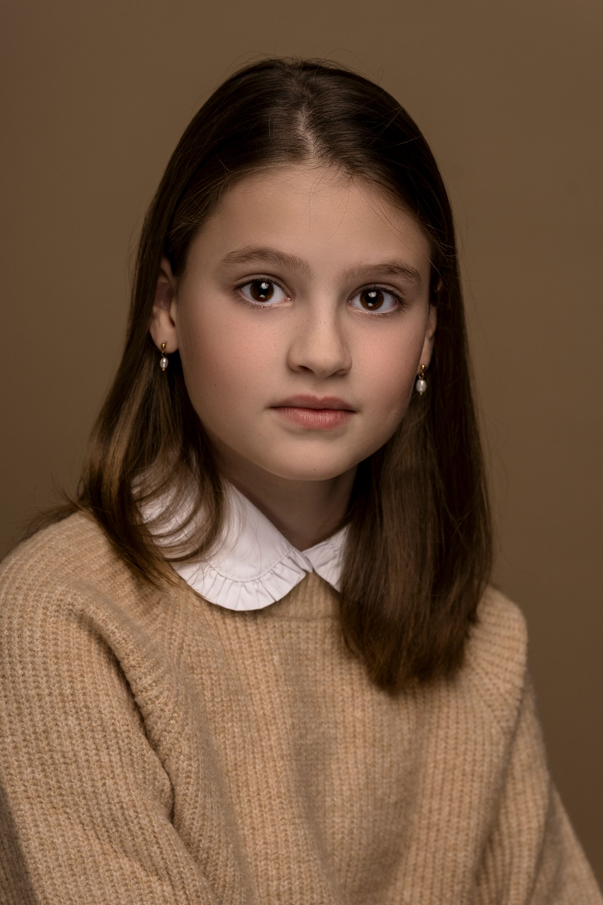 Meisje met lang blond haar, bleke huid, een lichtgekleurde trui, met haar hand op haar wang, kijkt naar de kijker.