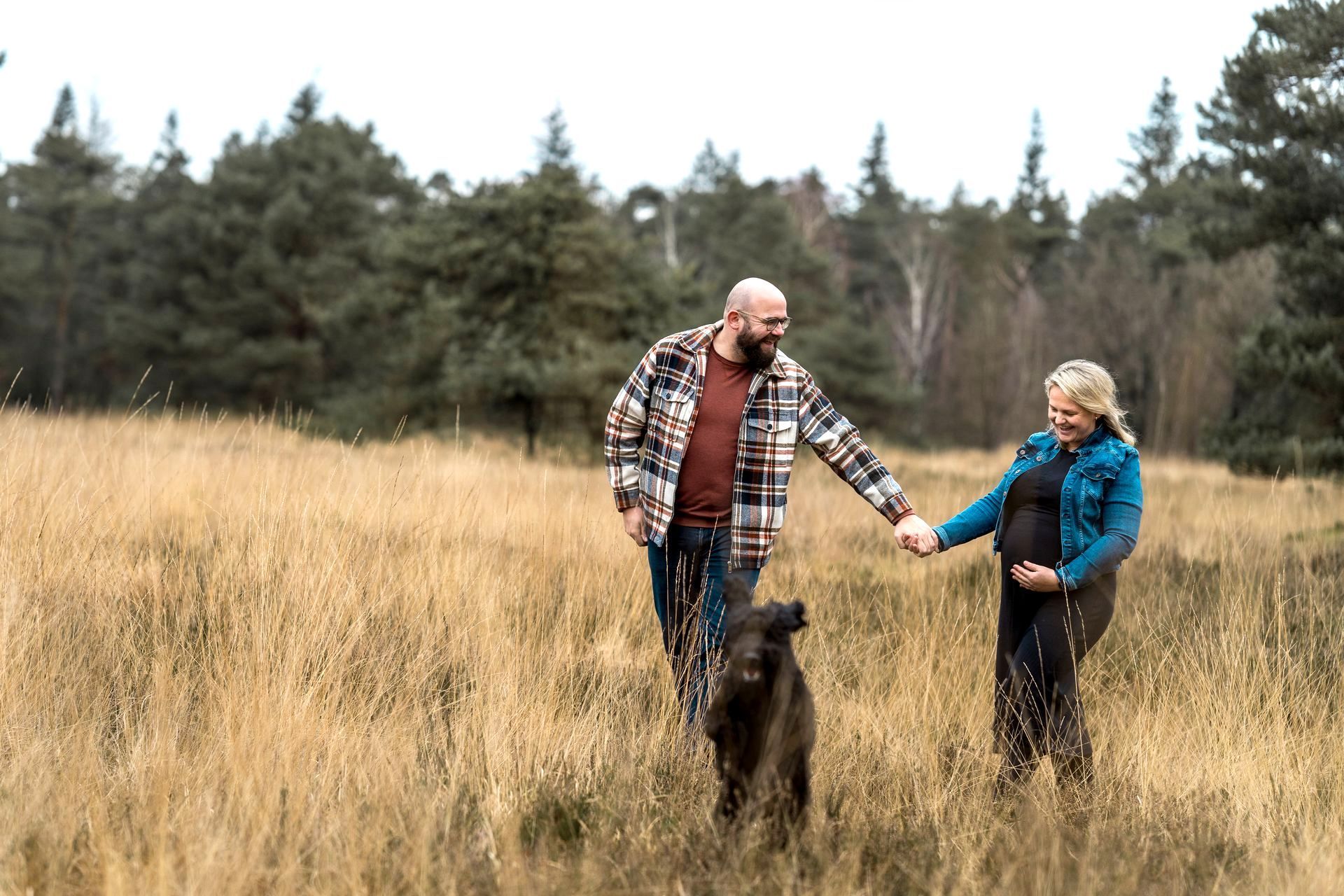 Een zwanger stel loopt hand in hand met hun hond door een veld, met een bos op de achtergrond.