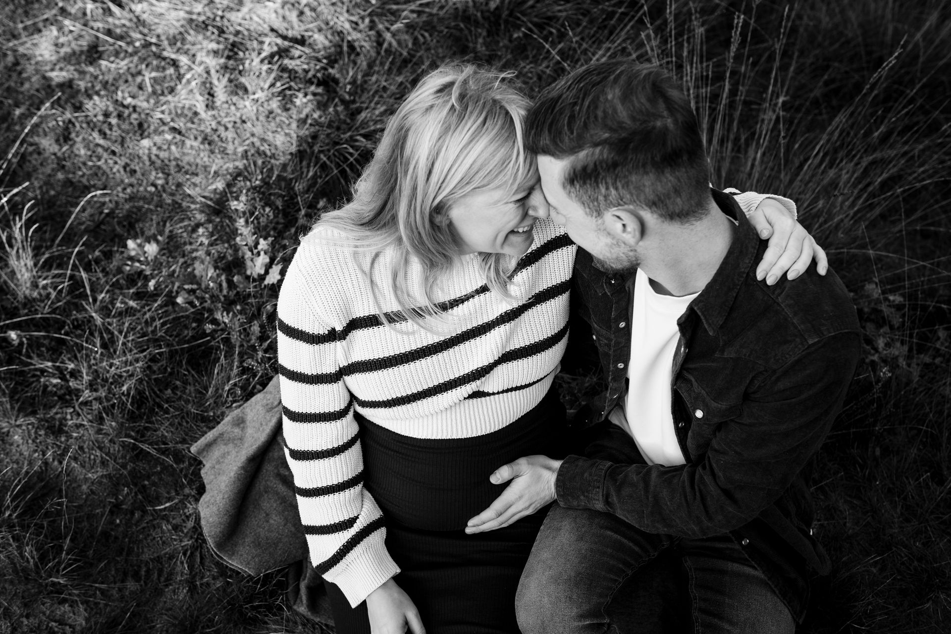 Een stel zit buiten en omhelst elkaar. De man raakt de zwangere buik van de vrouw aan, beiden glimlachen en raken elkaars hoofd aan. Zwart-wit.