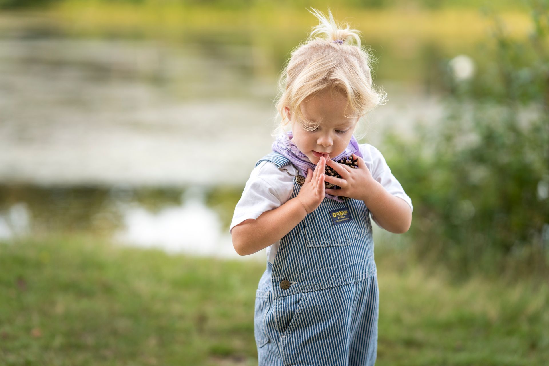 Een blond peutertje in een blauwe overall onderzoekt iets dat ze in kommetjesvormpjes vasthoudt, met gesloten ogen, buiten bij het water.